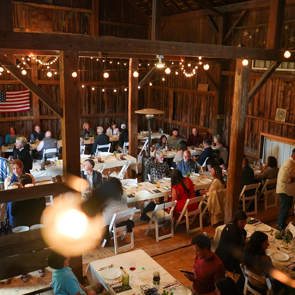 Group of people dining in a rustic barn setting with wooden tables and chairs.